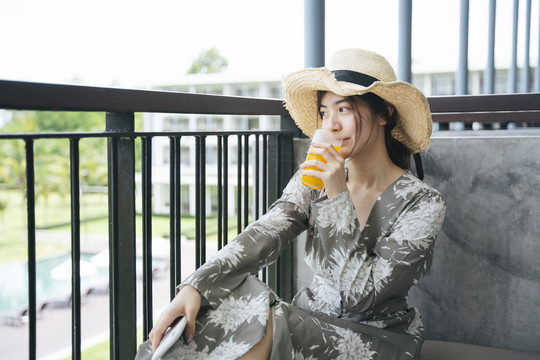 美丽的亚洲女游客戴着帽子在酒店阳台喝橙汁。