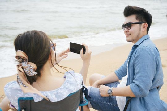 亚洲情侣旅行者坐在椅子上用智能手机在沙滩上拍照