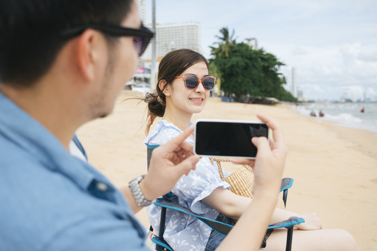 情侣旅行者坐在椅子上用智能手机在沙滩上拍照