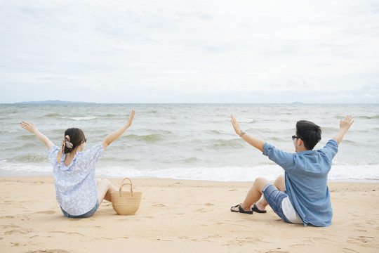 一对亚洲情侣坐在沙滩上，两只手举在空中。