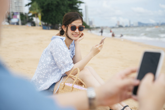 美丽的亚洲女人戴着墨镜用智能手机和情侣在沙滩上。