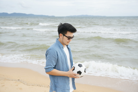 一个亚洲旅行者，穿着休闲衬衫，戴着墨镜，拿着足球在沙滩上散步。