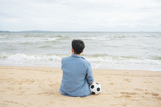 后视图-退休足球运动员拿着球坐在沙滩上。