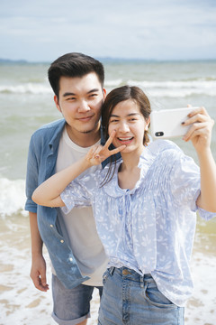 亚洲情侣旅行者在沙滩上用智能手机自拍。