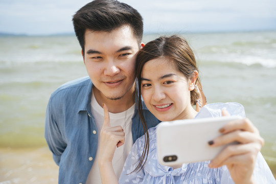 亚洲情侣旅行者在沙滩上用智能手机自拍。
