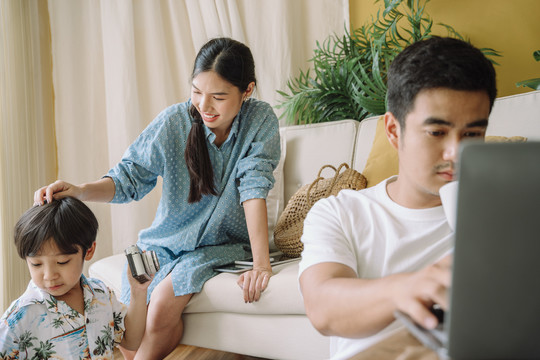 一个亚洲家庭在家共度时光。妈妈和儿子一起玩，而爸爸在笔记本电脑上工作。