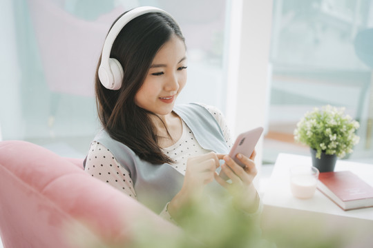 年轻漂亮的亚洲孕妇在家里用无线耳机和智能手机听音乐。