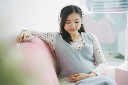 年轻漂亮的亚洲孕妇在家里用无线耳机听音乐。给未出生的孩子的旋律。