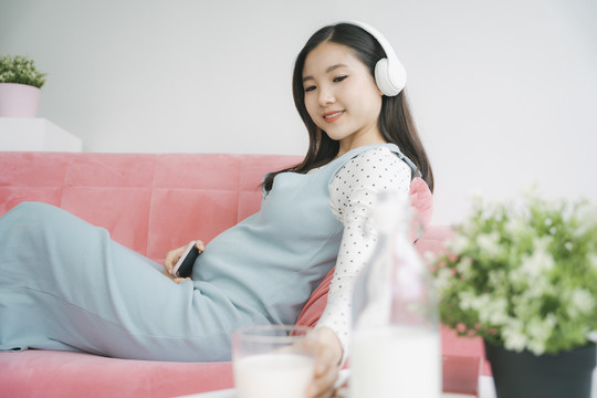 年轻漂亮的亚洲孕妇戴着无线耳机听音乐，一边在家里的沙发上休息一边抓着一杯牛奶。