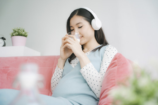 音乐爱好者怀孕的亚洲妈妈在客厅用无线耳机听音乐，喝一杯牛奶。