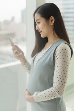 侧视图-年轻美丽的泰国亚裔孕妇使用智能手机查看社交媒体。