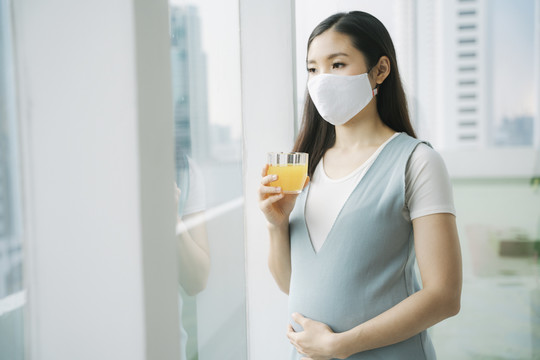 美丽的亚洲孕妇戴着棉质口罩，在家里捧着一杯橙汁。健康饮料。