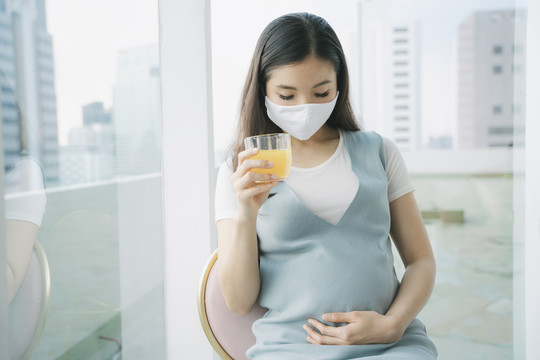 美丽的亚洲孕妇戴着棉质口罩坐在椅子上，手里拿着一杯橙汁。在家里隔离。