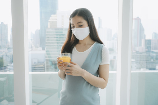 美丽的亚洲孕妇戴着棉质口罩拿着橙汁在家。在家里隔离。