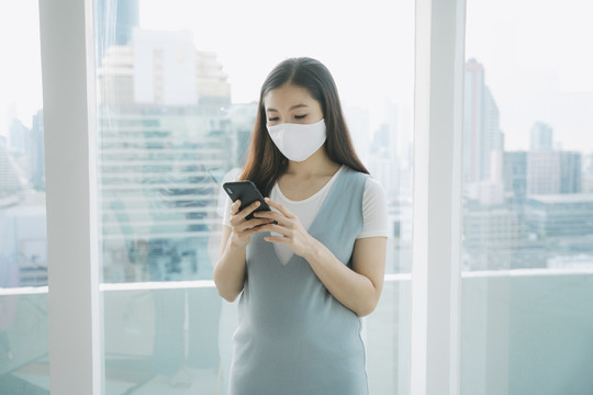 美丽的亚洲孕妇戴着口罩在家使用智能手机。检疫。