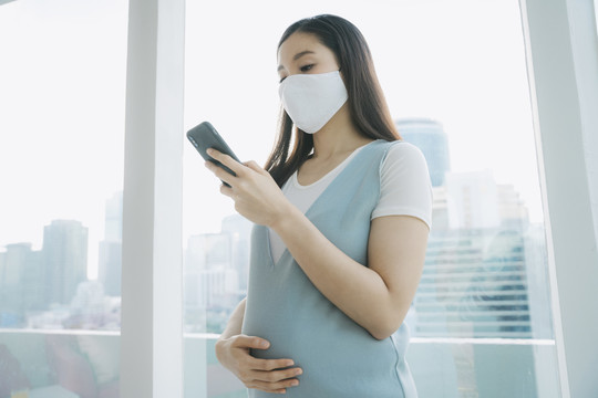 美丽的亚洲孕妇戴着棉质口罩在家使用智能手机。在家里隔离。