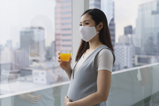 美丽的亚洲孕妇戴着棉质口罩拿着橙汁望着窗外。