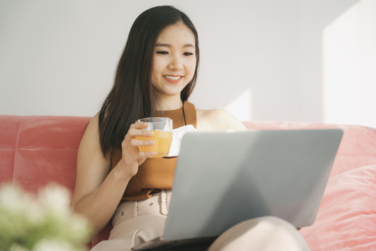 美丽的亚泰女商人在家拿着一杯橙汁在笔记本电脑上工作。