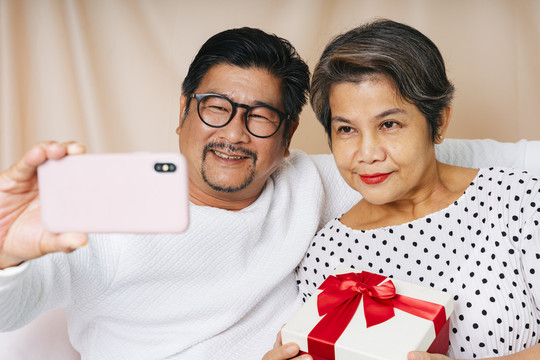 亚洲老年夫妇在生日派对上用智能手机自拍。妻子拿着礼物盒。