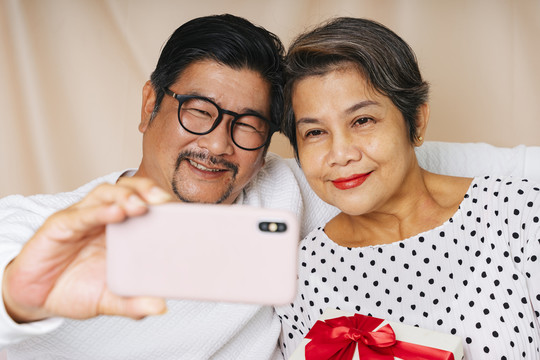 亚洲老年夫妇在生日派对上用智能手机自拍。妻子拿着礼物盒。