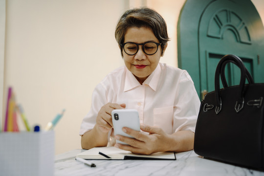 亚洲老年妇女在餐桌上用智能手机戴眼镜。