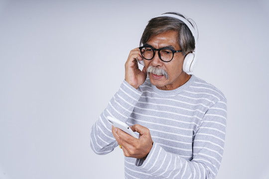 照片中的老人在白色背景下用无线耳机和智能手机听音乐。