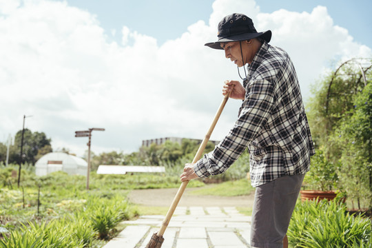 在一个农场里，一位亚洲老年农民用扫帚杆打扫花园。