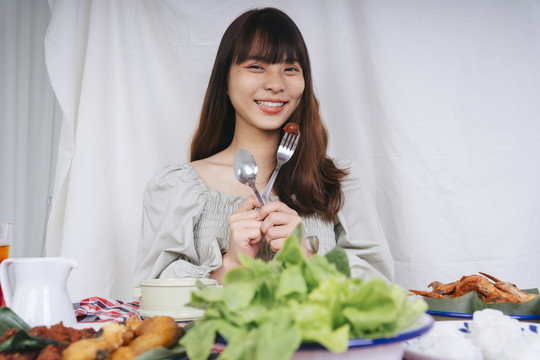 亚洲妇女拿着勺子和叉子在家里吃是一种食物的肖像。