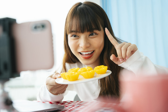 年轻的亚洲黑发女子在智能手机摄像头前拿着金色蛋糕做泰国甜点食品评论。