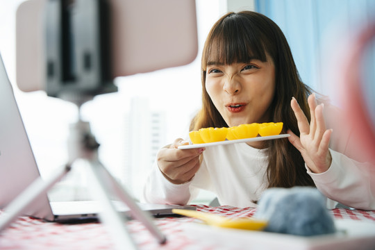 年轻的亚洲黑发女子在智能手机摄像头前拿着金色蛋糕做泰国甜点食品评论。