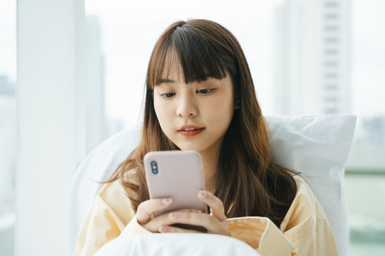 年轻的亚洲黑发女子早上在床上使用智能手机。