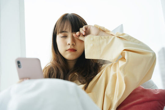 昏昏欲睡的亚洲黑发女青年早上醒来躺在床上用智能手机。