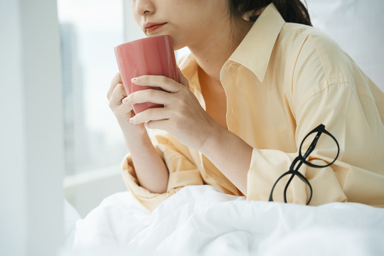 侧视图-年轻的亚洲女人早上在床上拿着一杯咖啡。