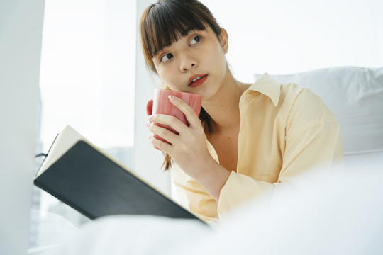 年轻的亚洲黑发女人早上在床上看书喝咖啡。