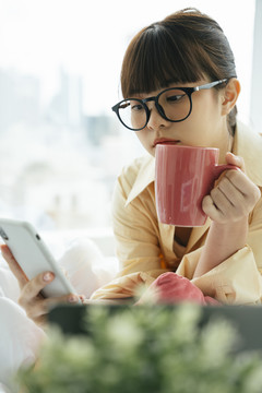 年轻的亚洲黑发女子戴着眼镜，手里拿着一杯咖啡和智能手机。