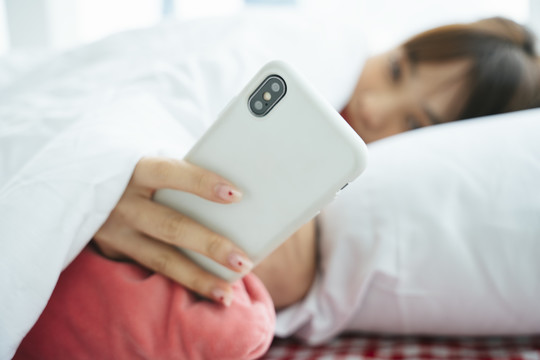 年轻的亚洲黑发女子早上躺在床上用智能手机。