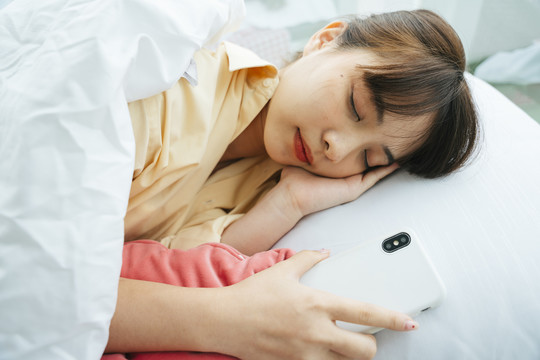 年轻的亚洲黑发女子抱着智能手机睡在床上。