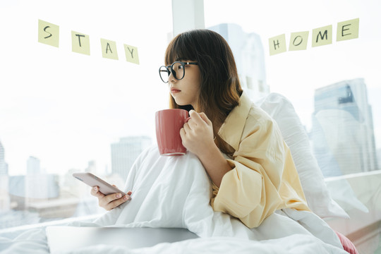 年轻的亚洲黑发女子戴着眼镜拿着咖啡和智能手机躺在卧室里看着窗外。
