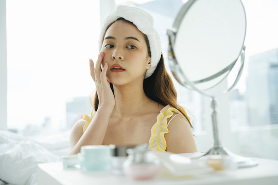 年轻的亚洲黑发女子在镜子前使用面部护肤品。