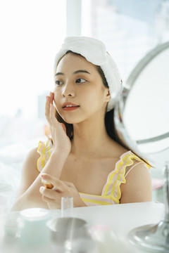 年轻的亚洲黑发女性在镜子前使用面部美容护肤品。