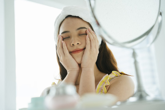 年轻的亚洲黑发女人在镜子前用美容护肤品闭上眼睛。