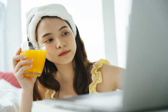 年轻的亚洲黑发女子拿着一杯橙汁用笔记本电脑。