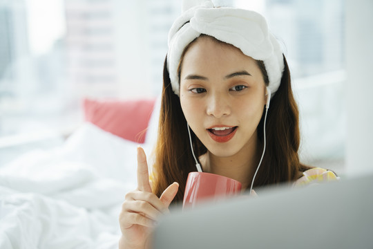 年轻的亚洲黑发女子戴着白色的头巾捧着一杯咖啡用耳机听音乐和笔记本电脑上的人交谈。