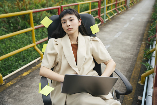 在户外的一座桥上，一位穿着正式衬衫的亚洲女商人坐在椅子上使用笔记本电脑。