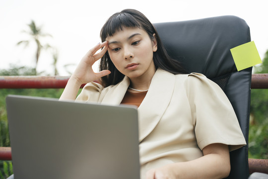 体贴的亚洲女商人穿着正式的衬衫坐在椅子上，在户外使用笔记本电脑。