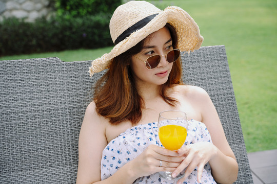 美丽的亚洲女人戴着帽子和太阳镜在沙滩椅上放着橙汁放松。