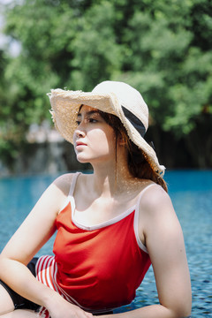美丽的亚洲女游客穿着红衬衫，戴着草帽，在室外的游泳池里放松。