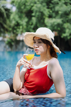美丽的亚洲女游客穿着红衬衫，戴着草帽，在游泳池里喝着橙汁放松。暑假概念。