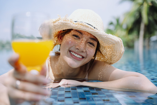 美丽的亚洲女游客戴着帽子捧着一杯橙汁在游泳池里放松。