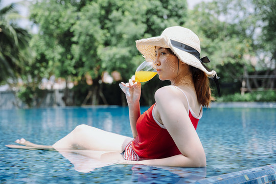 侧视图-美丽的亚洲女游客穿着红色泳衣和帽子在游泳池喝橙汁。